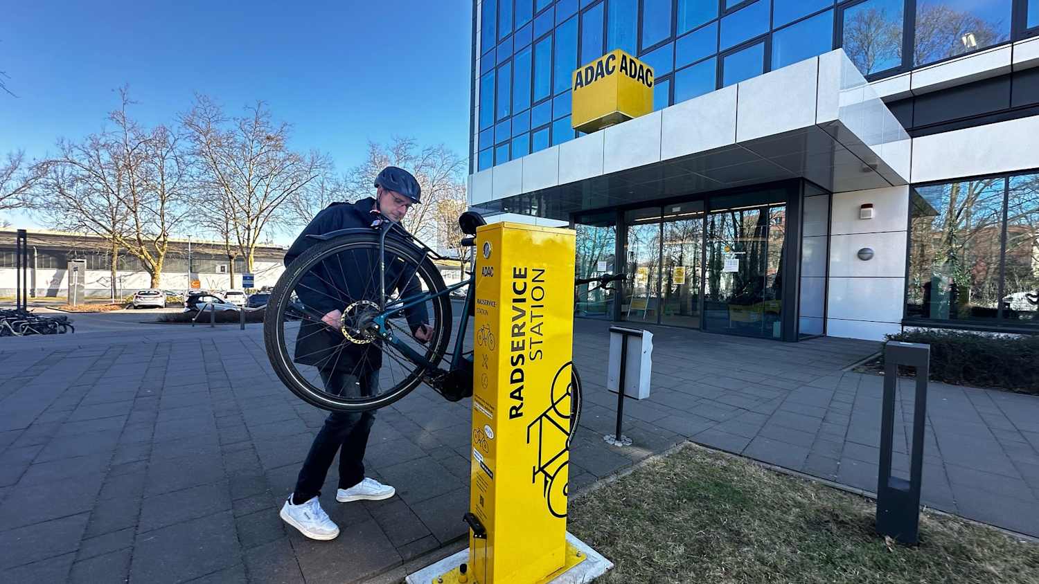 Ein Mann hängt ein Fahrrad an eine gelbe Säule mit der Aufschrift Radservicestation. Im Hintergrund ist ein verglastes Hochhaus mit ADAC Logo zu sehen.