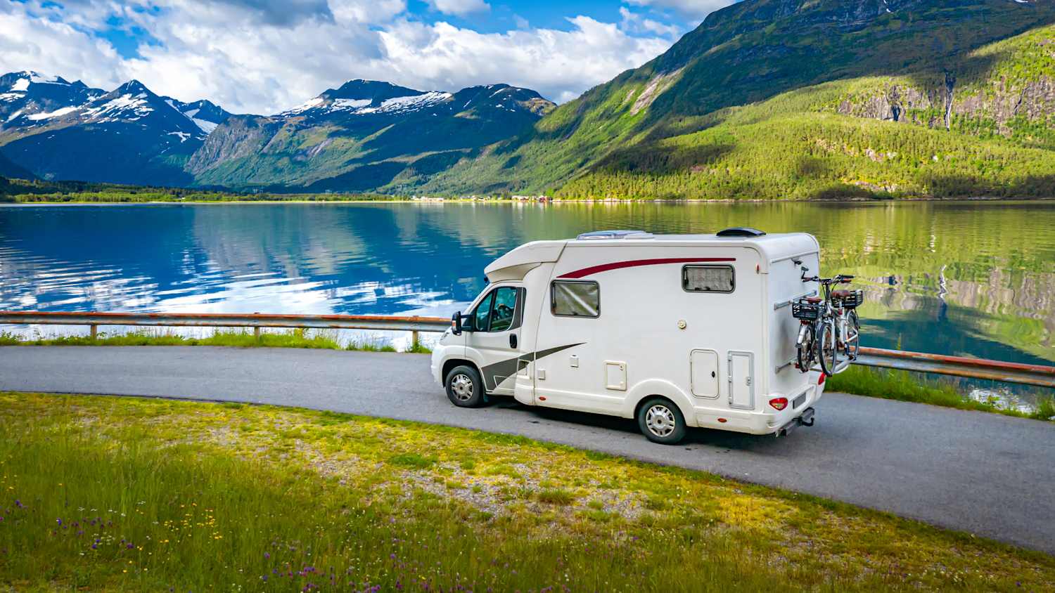 Ein Wohnwagen fährt entlang einer Straße am See. Im Hintergrund sind Berge zu sehen.