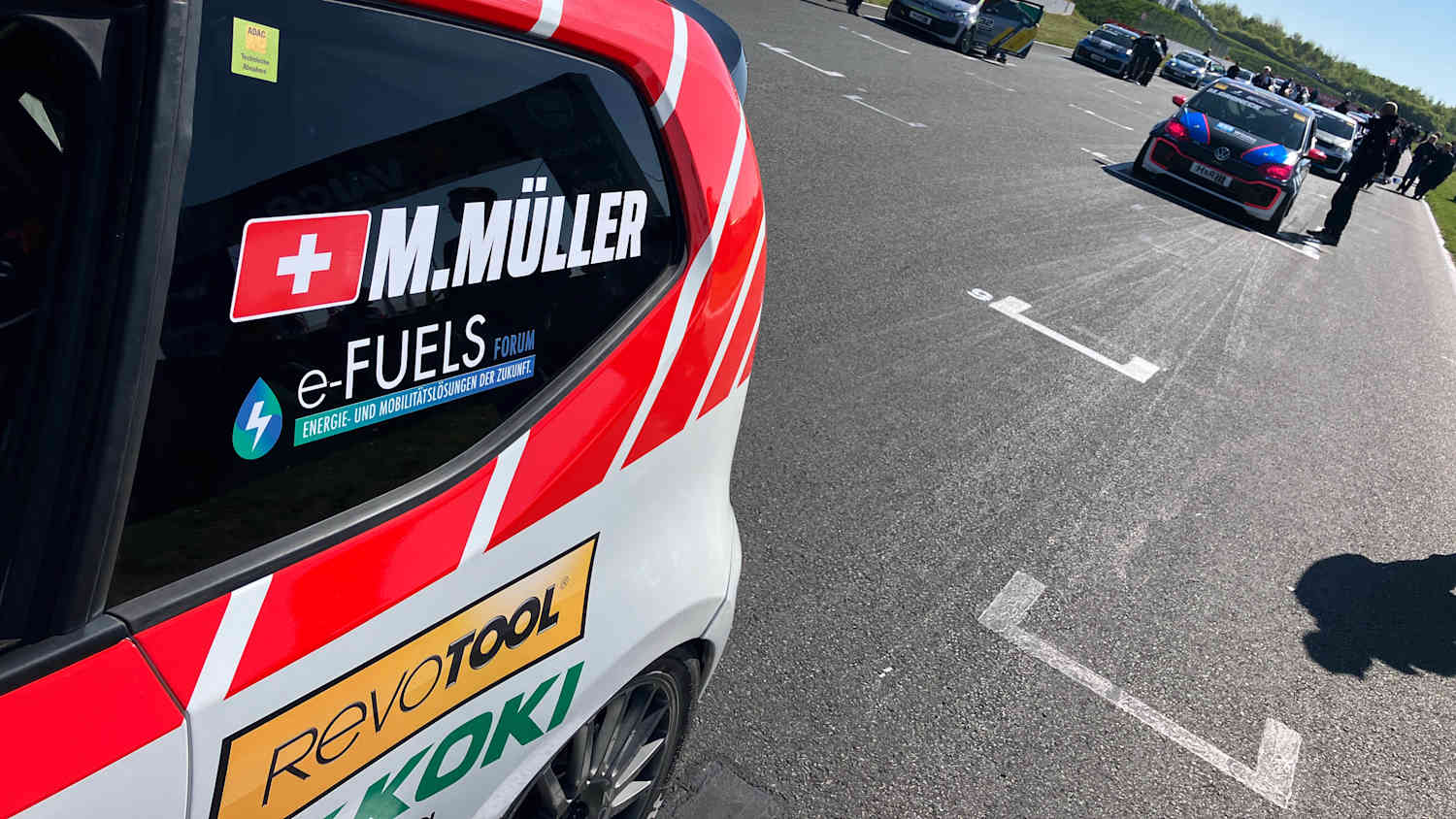 Eine Rennstrecke mit mehreren Autos unter blauem Himmel ist zu sehen. Links in Vordergrund steht ein Auto mit einem eFuels Forum Sticker.