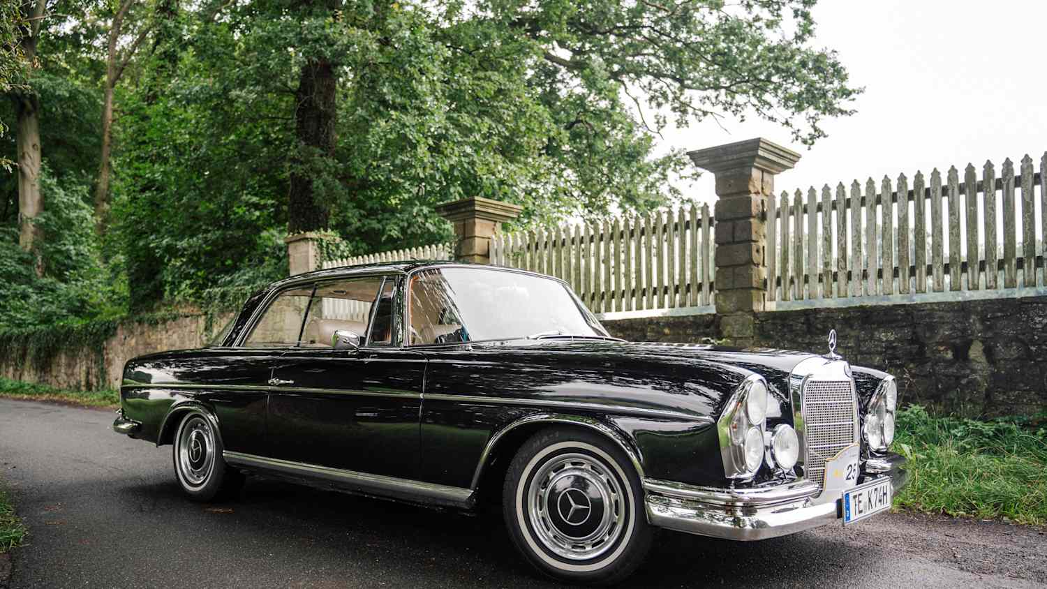 Ein schwarzer Oldtimer von Mercedes steht auf einer Straße. Im Hintergrund sieht man einen Zaun und einen Baum.