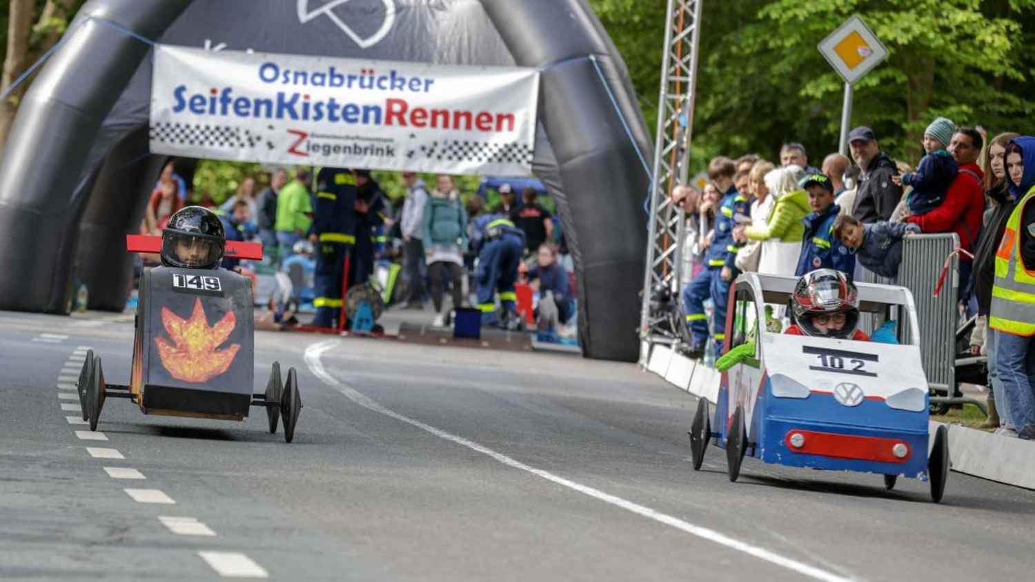 Auf einer Rennstrecke fahren zwei Seifenkisten nebeneinander. Im Hintergrund ist das Publikum zu sehen.