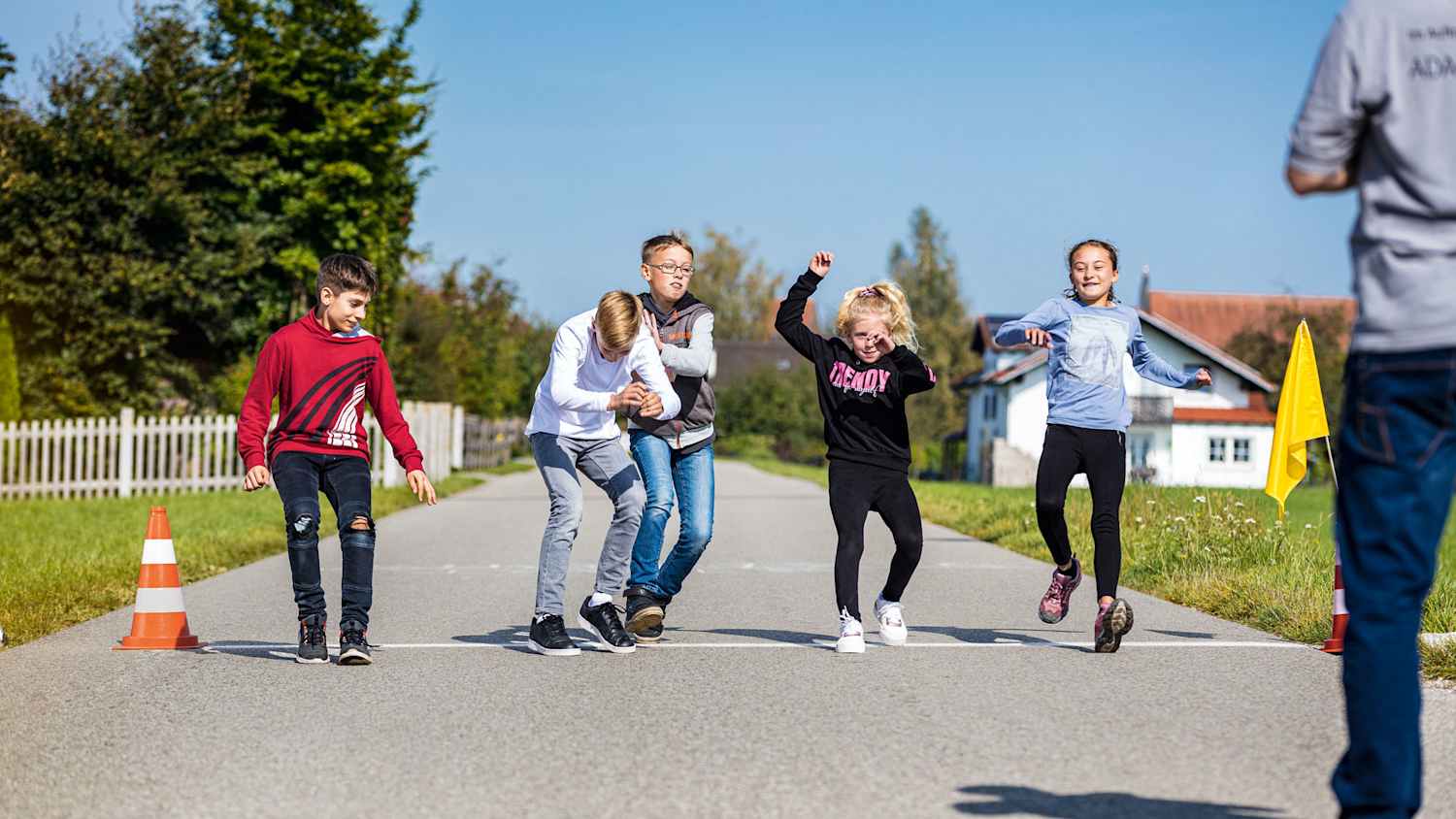 Fünf Kinder stehen auf einer Straße nebeneinander an einer weißen Linie. Die Linie ist auf den Boden gemalt und an beiden Enden der Linie steht ein Verkehrskegel. Rechts vor den Kindern steht ein Trainer.