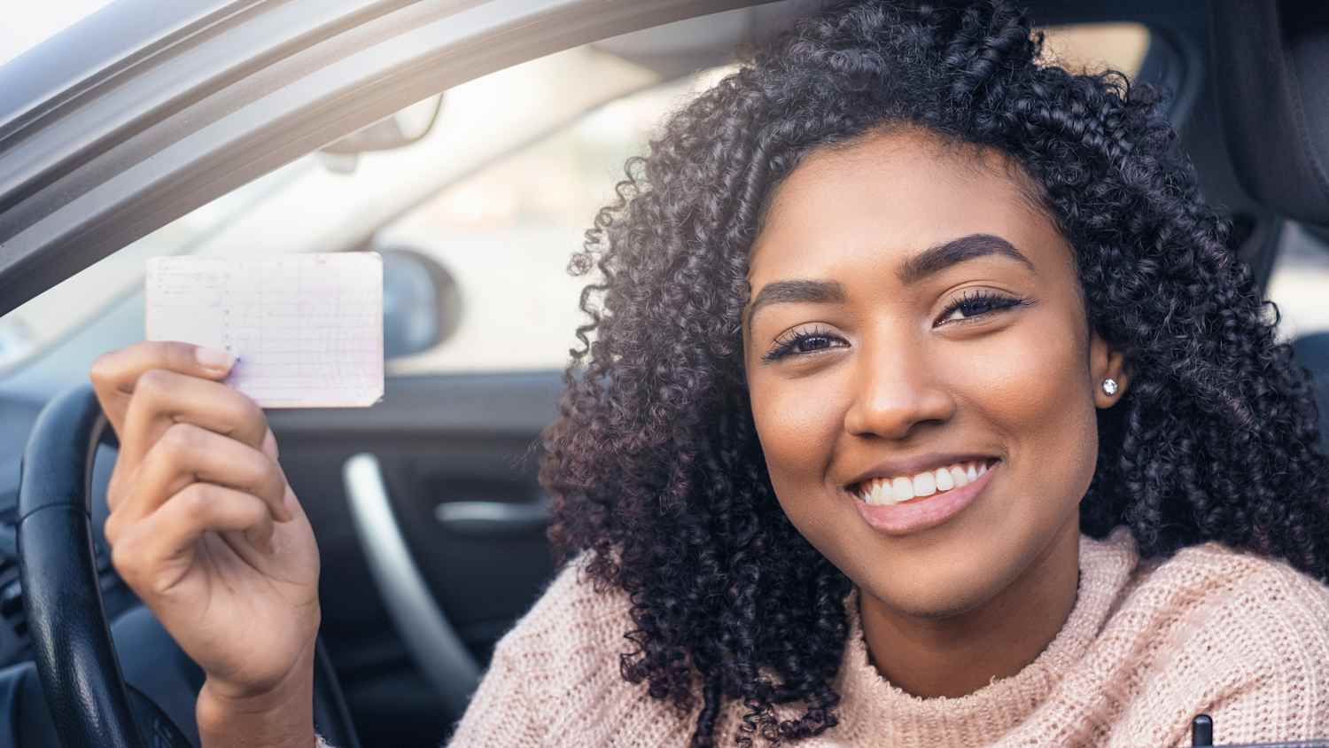 Eine lächelnde Frau sitzt im Auto und hält ihren Führerschein in die Kamera.