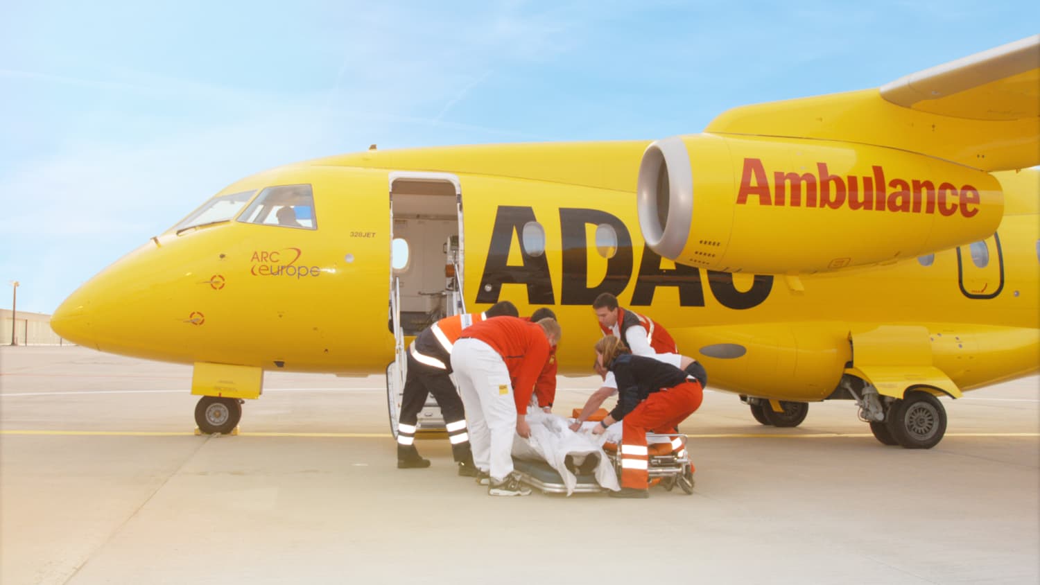 Ein ADAC-Ambulance-Flugzeug steht auf einem Rollfeld. Die Kabinentür steht offen, da ein Patient durch das Rettungspersonal ins Flugzeug gebracht werden soll für einen Krankenrücktransport.