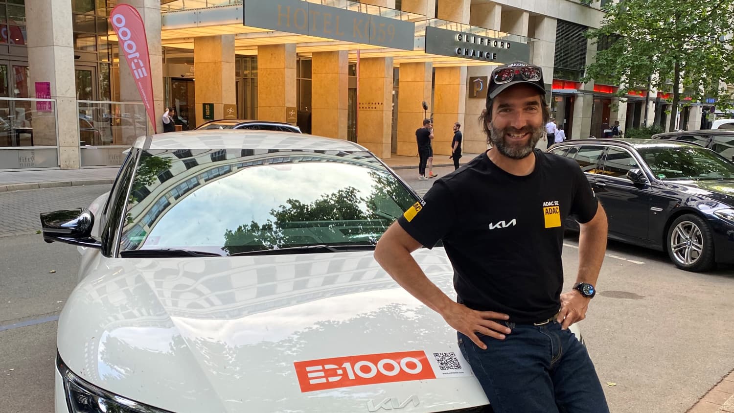 Mann in ADAC T-shirt sitzt auf Motorhaube eines weißen Kia EV6, der durch Kia Leasing jetzt beim ADAC verfügbar ist.