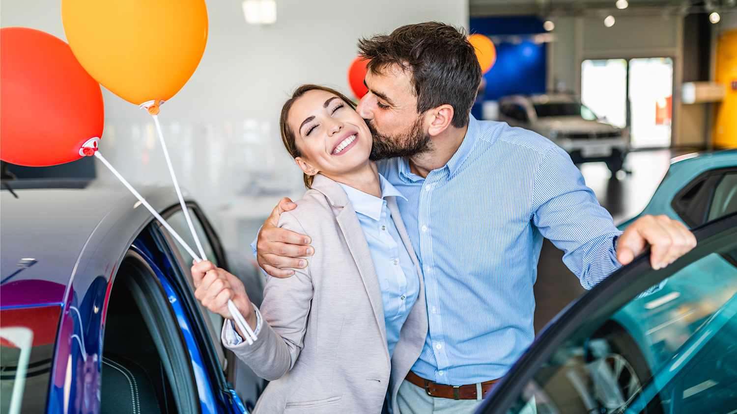 Pärchen steht vor dem neu gekauften Auto und die Frau hält Luftballons in der Hand