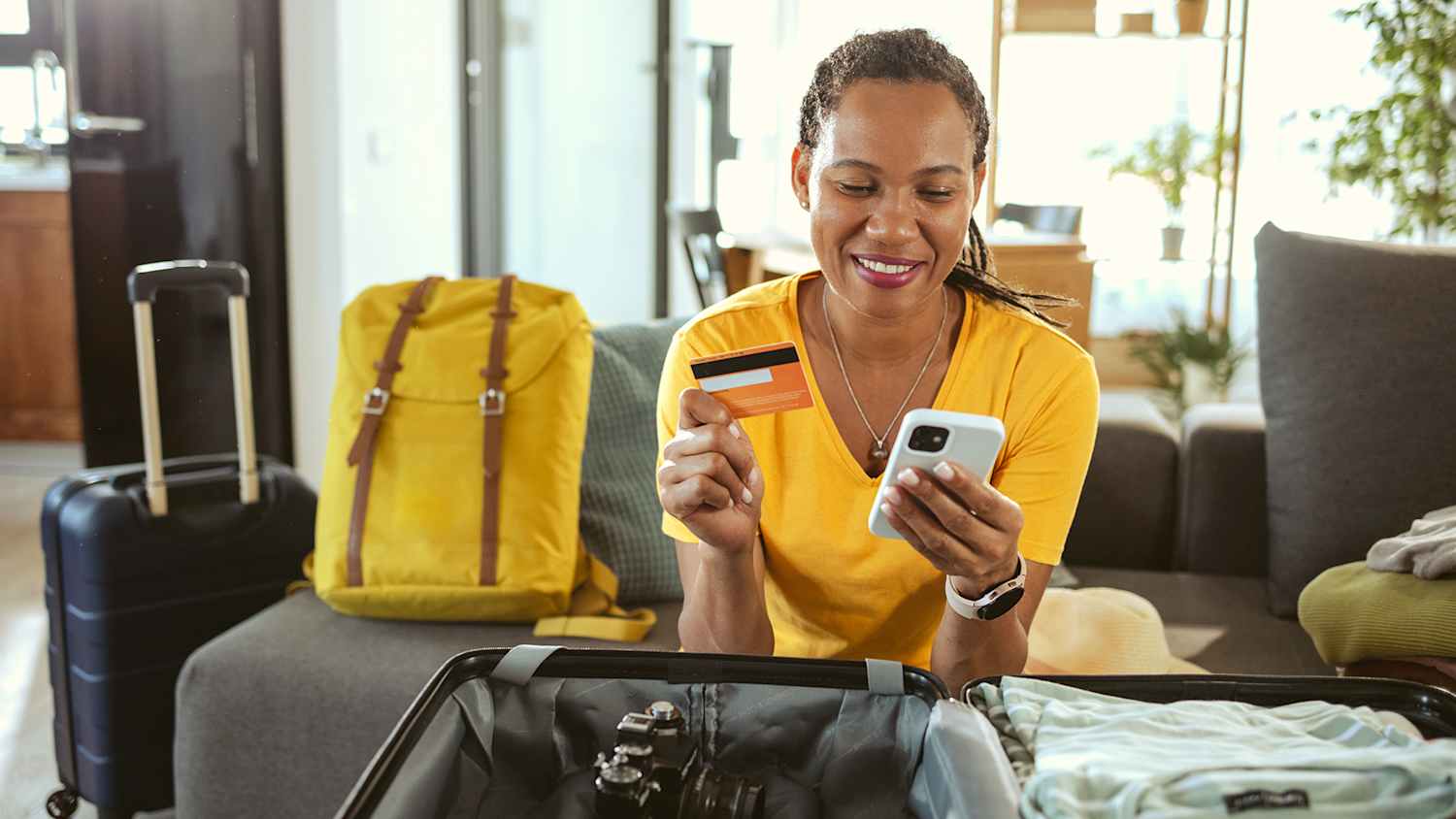 Eine Frau sitzt lächelnd auf einer Coach neben gepackten Taschen und hat eine kreditkarte für die reise im Gepäck