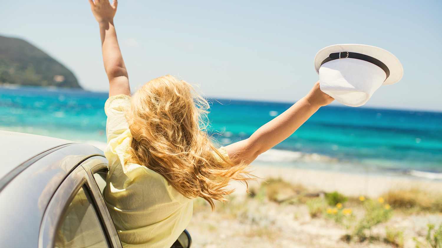 Eine Frau streckt sich am Strand aus dem Fenster ihres Autos, denn laut einer ADAC Umfrage fahren Menschen weiterhin am liebsten mit dem Auto in den Urlaub