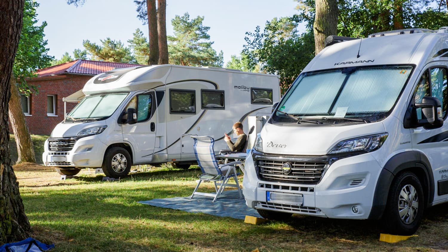 Zwei weiße Wohnmobile stehen auf einem Campingplatz nebeneinander, ein Mann sitzt auf einem Stuhl dazwischen und sieht in seinem Handy nach Wohnmobile zu kaufen