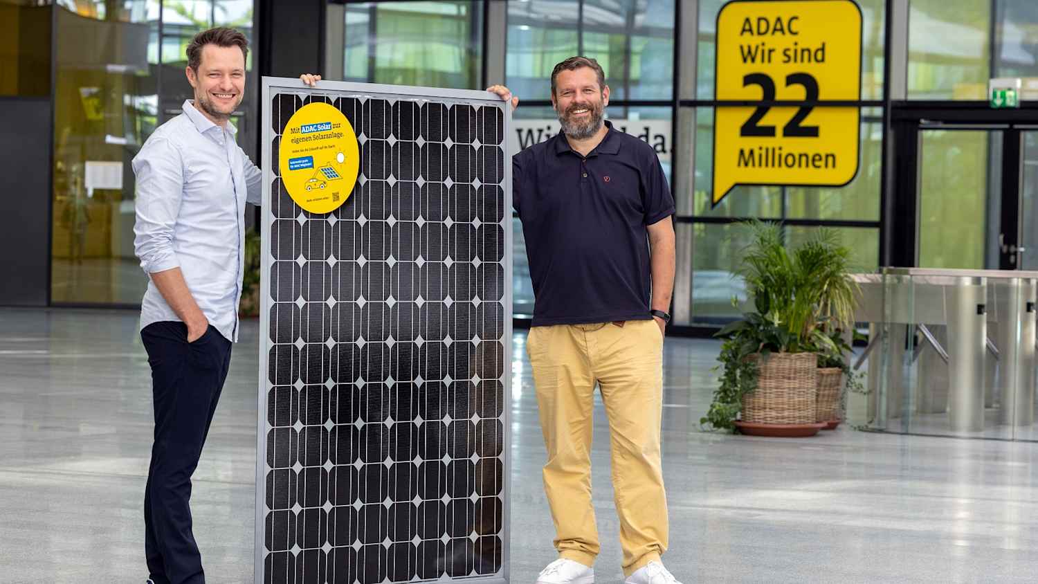 Zwei Männer halten ein Solarpanel aufrecht in dem Foyer eines Gebäudes und verdeutlichen so die Zusammenarbeit zwischen adac und otovo.