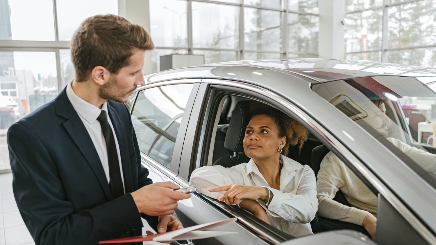 Ein Mann und eine Frau sitzen in einem grauen Auto im Autosalon und erhalten den Schlüssel von einem Autohändler, nachdem sie die Frage leasingauto abholen oder liefern geklärt haben.