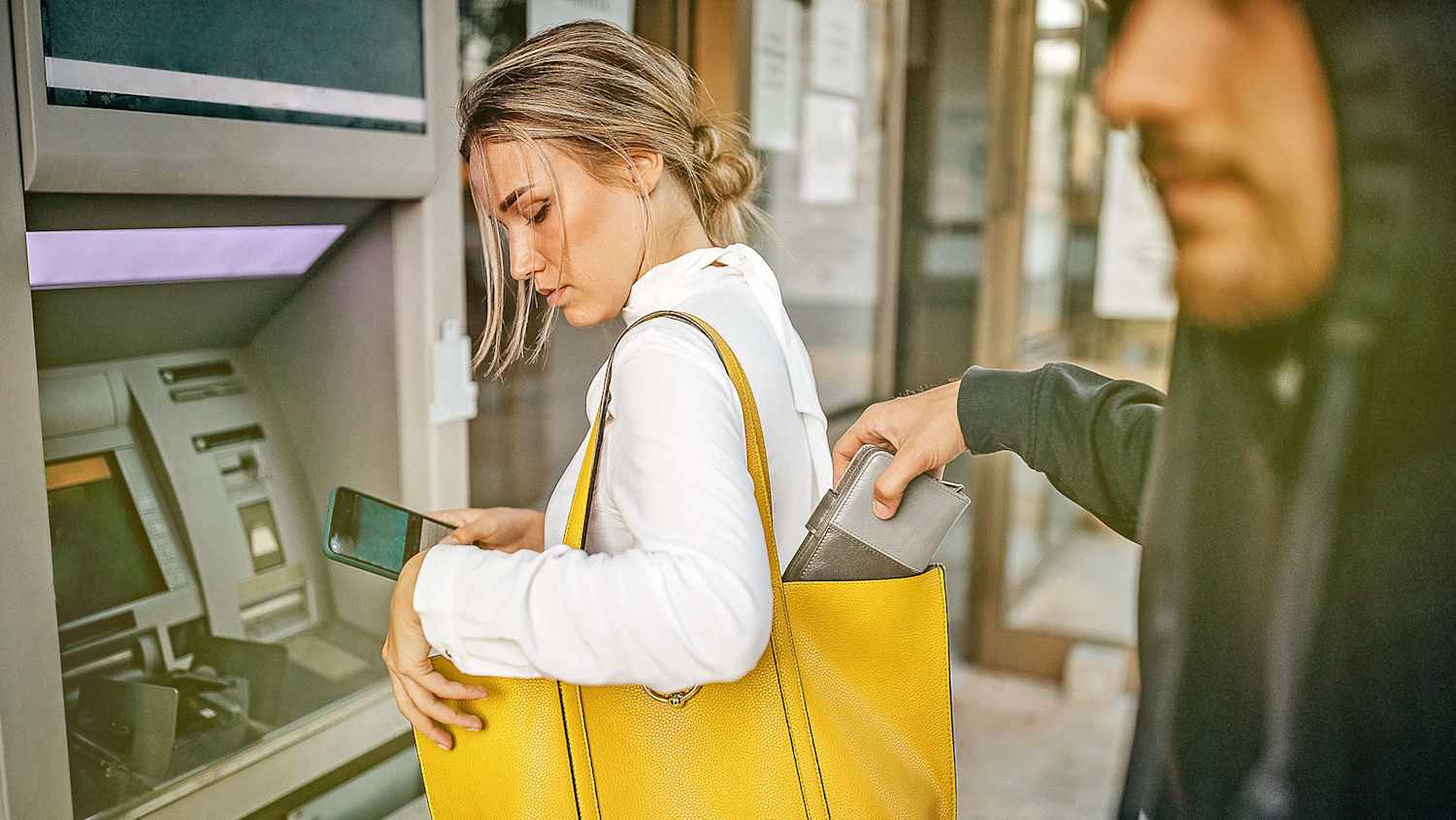 Frau mit Smartphone steht am Geldautomaten, Dieb stiehlt Geldbeutel aus Handtasche.