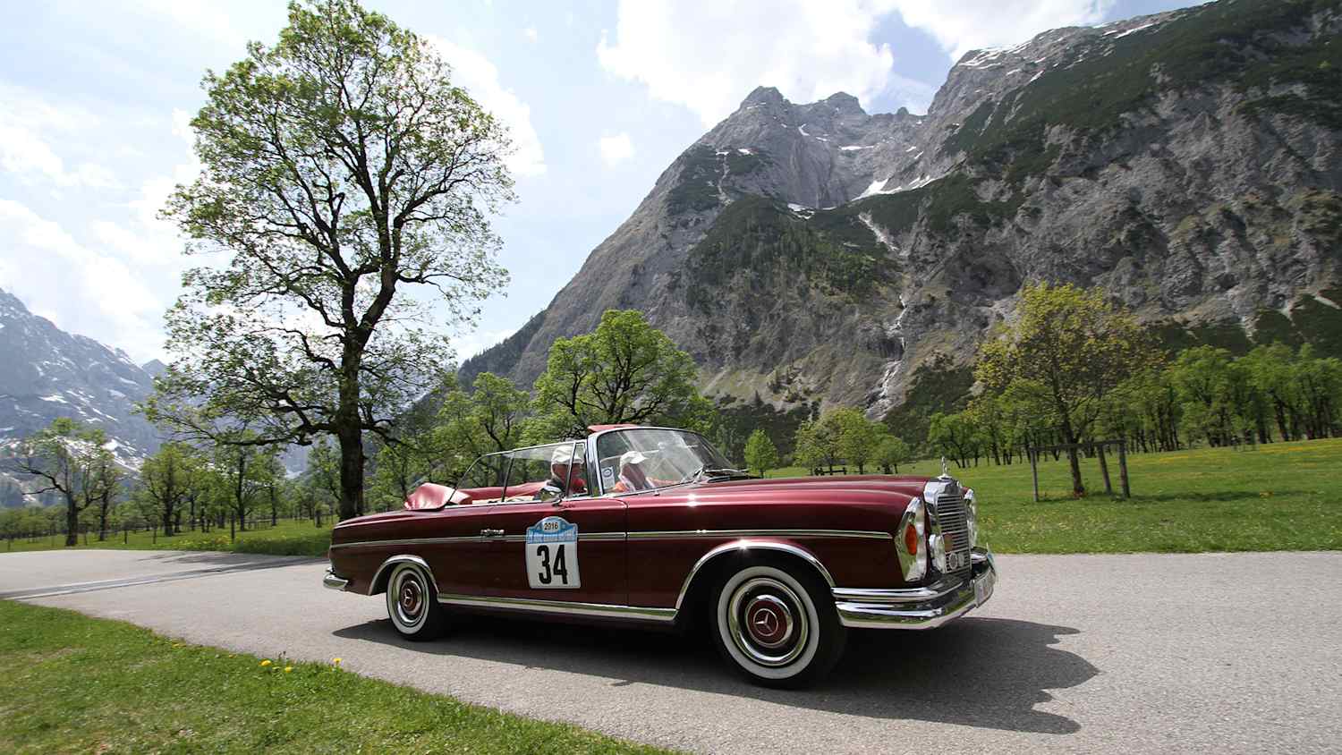 Zwei Männer fahren in einem roten Oldtimer eine Straße neben den Bergen entlang und müssen sich keine Gedanken über eine Oldtimerversicherung machen