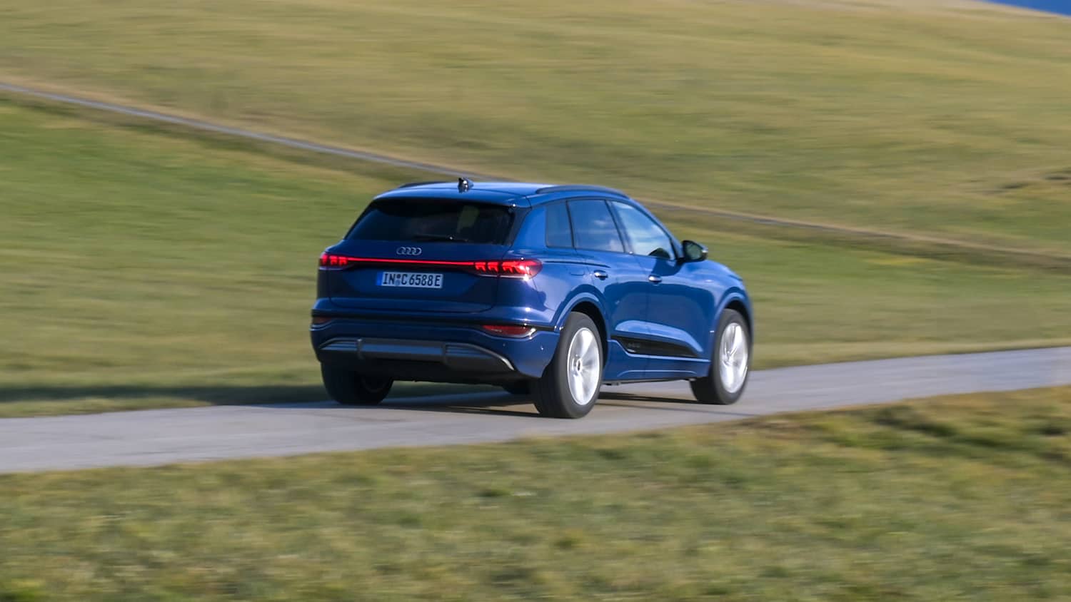 Audi Q6 e-tron fährt über eine Straße, Heckansicht mit Bäumen und Himmel im Hintergrund.