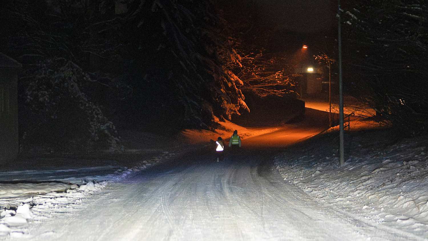 Zwei Kinder mit Warnwesten laufen auf einer schneebedeckten Straße. Eine der Warnwesten leuchtet, die andere nicht