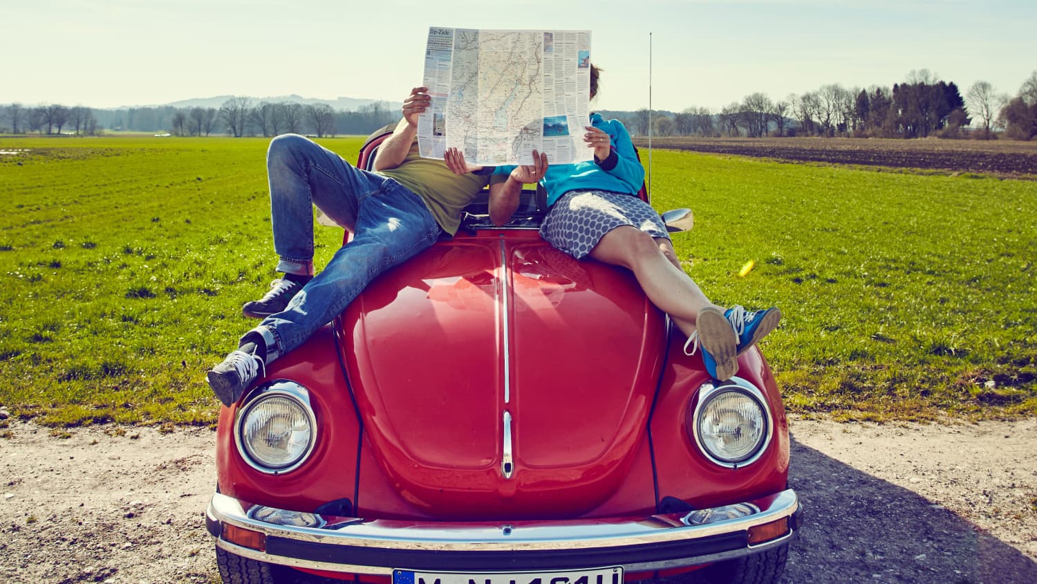 Ein Paar liegt auf einem roten VW Käfer und studiert eine Landkarte