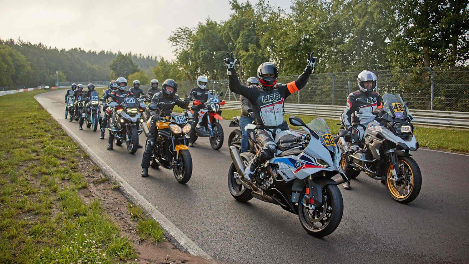 Eine Gruppe von Motorradfahrern fährt auf einer Straße, umgeben von grüner Landschaft und Bäumen.