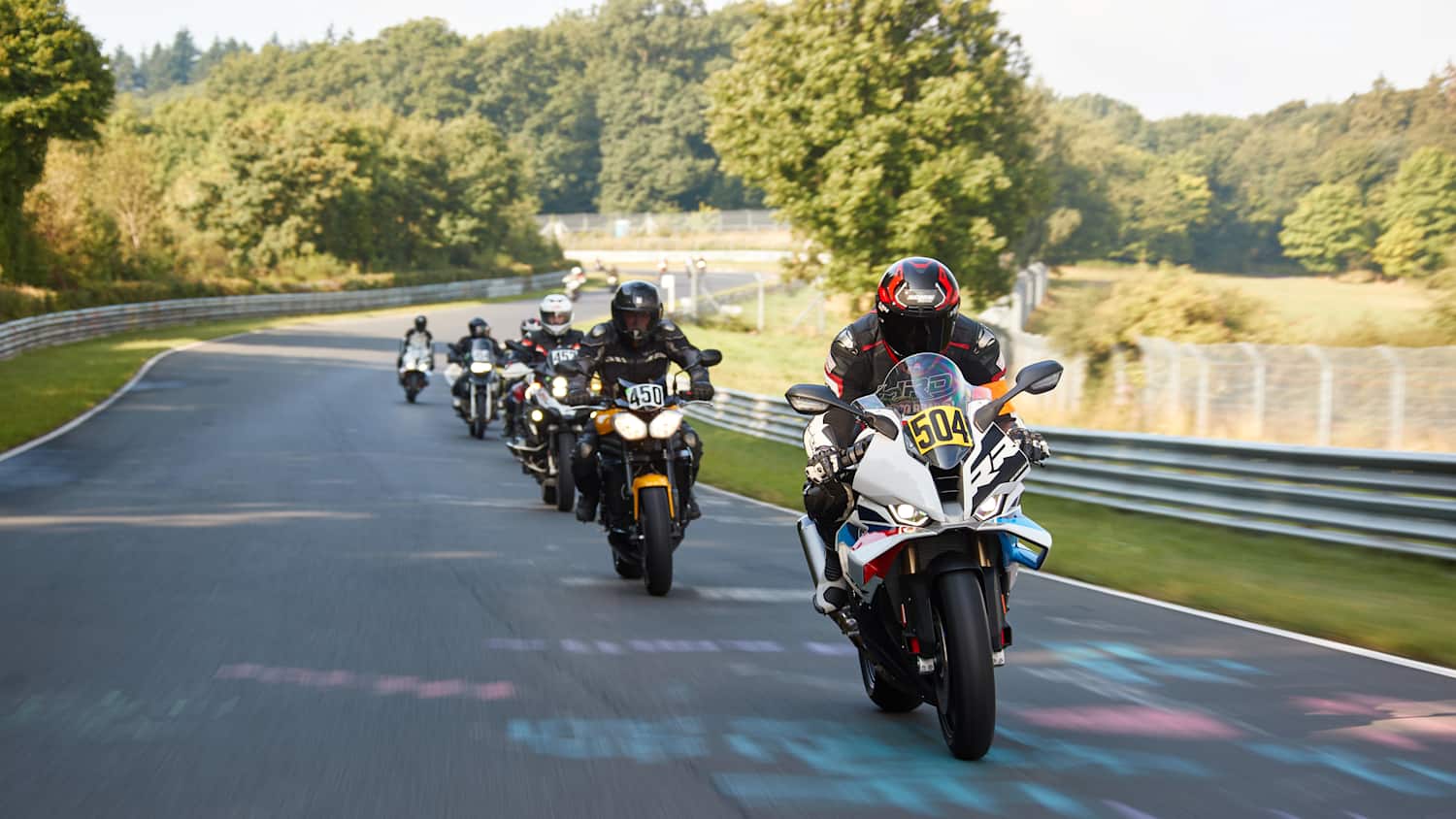 Eine Gruppe von Motorradfahrern fährt auf einer Straße, umgeben von Bäumen und grüner Landschaft.