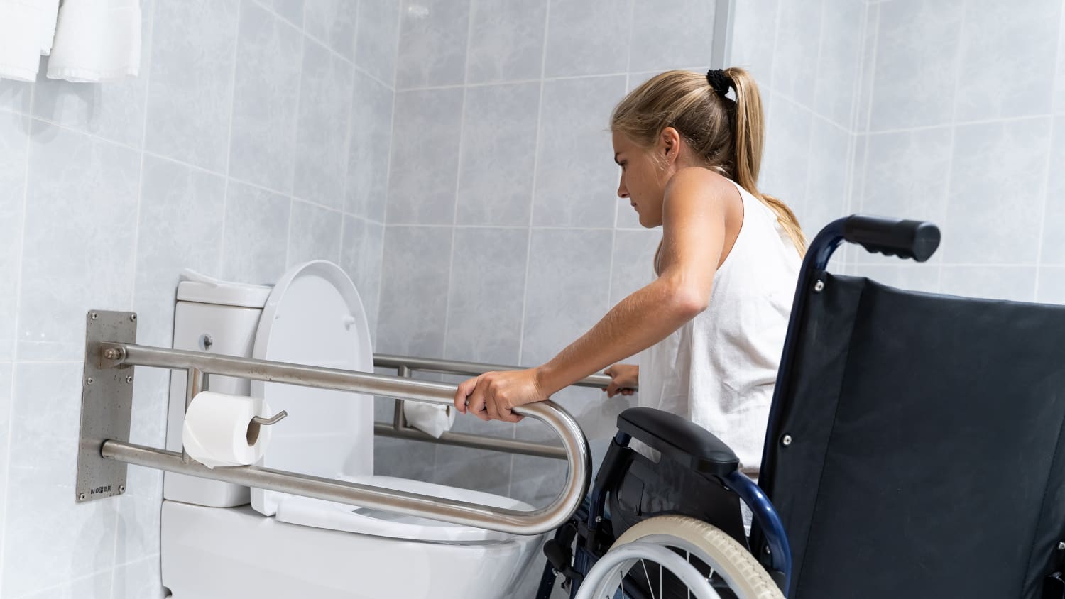 Eine Frau hält sich am Toilettengeländer fest und versucht, mit den Armen aus dem Rollstuhl aufzustehen, um auf die Toilette zu gehen