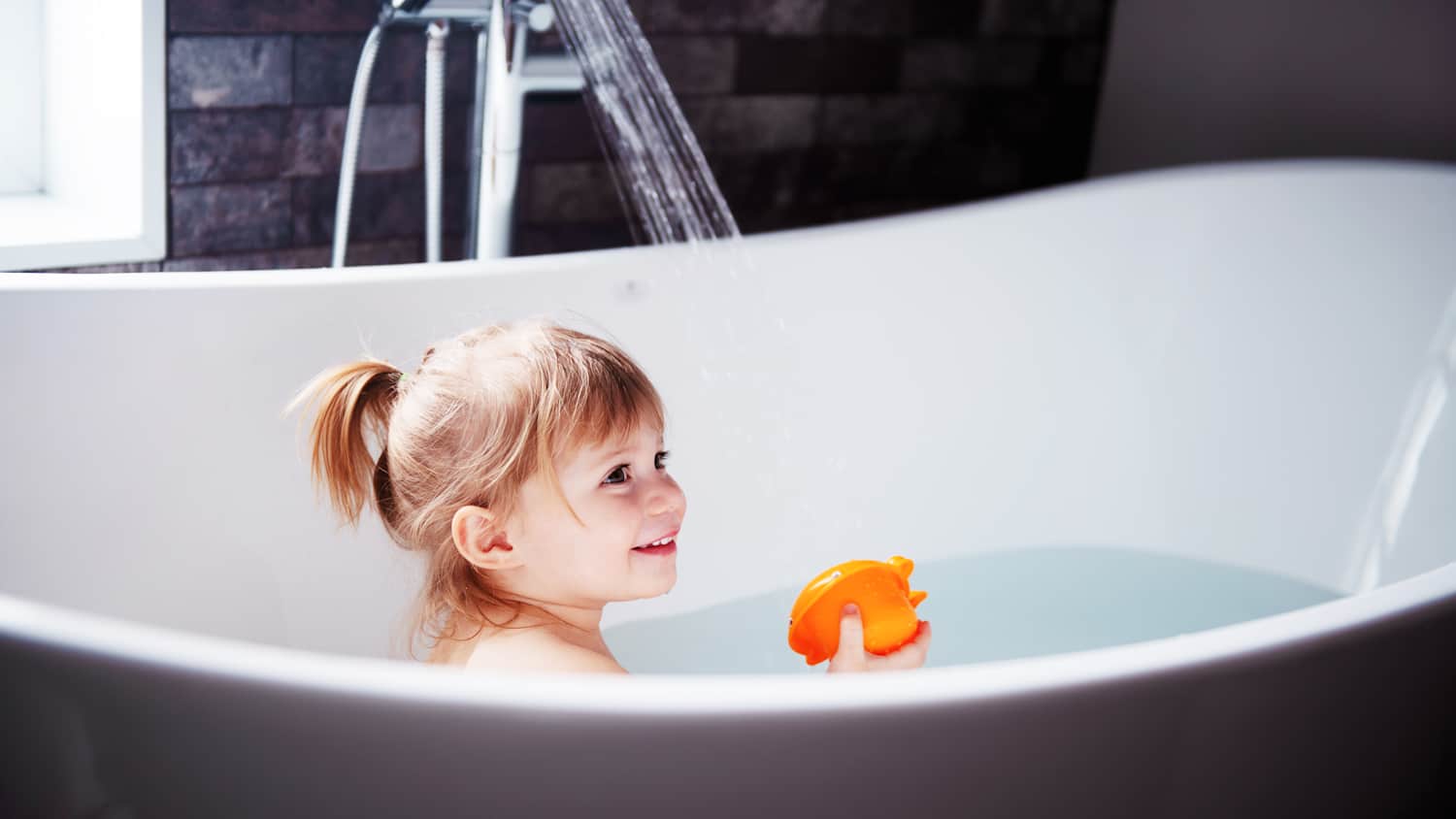 Ein kleines Mädchen welches mit einem Schwimmtier in der Hand, in der Badewanne sitzt & Freude hat