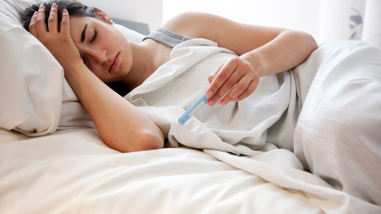 Eine Frau liegt mit Fieber im Bett und misst ihre Temperatur