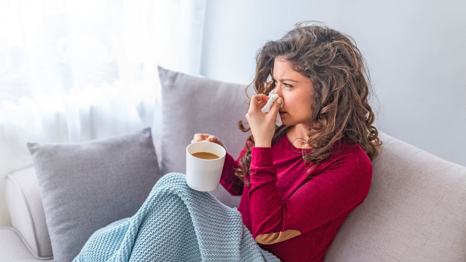 Eine Frau sitzt auf einem Sofa mit einer Decke über den Beinen und einer Tasse in der Hand und putzt sich die Nase