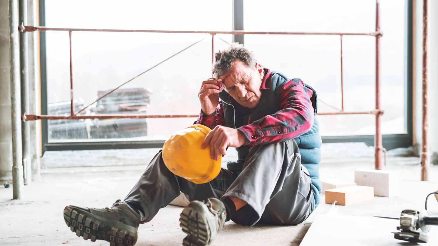 Ein Mann hält sich nach einem Unfall auf einer Baustelle ein Tuch an die blutende Stirn in der andern Hand hält er einen gelben Bauhelm