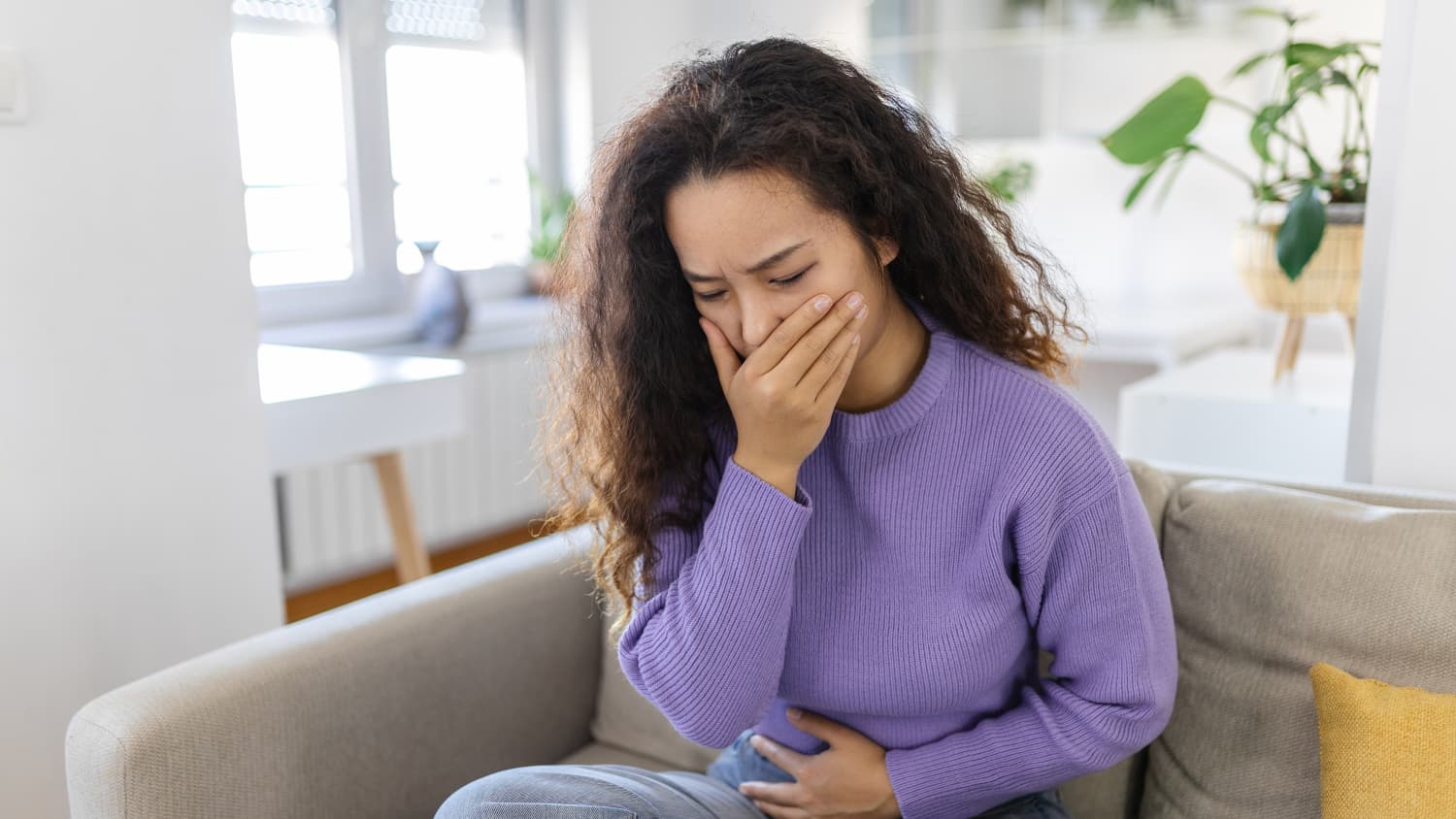 Eine Frau, in lila Pullover, auf einer Couch hält sich wegen Übelkeit die Hand vor den Mund