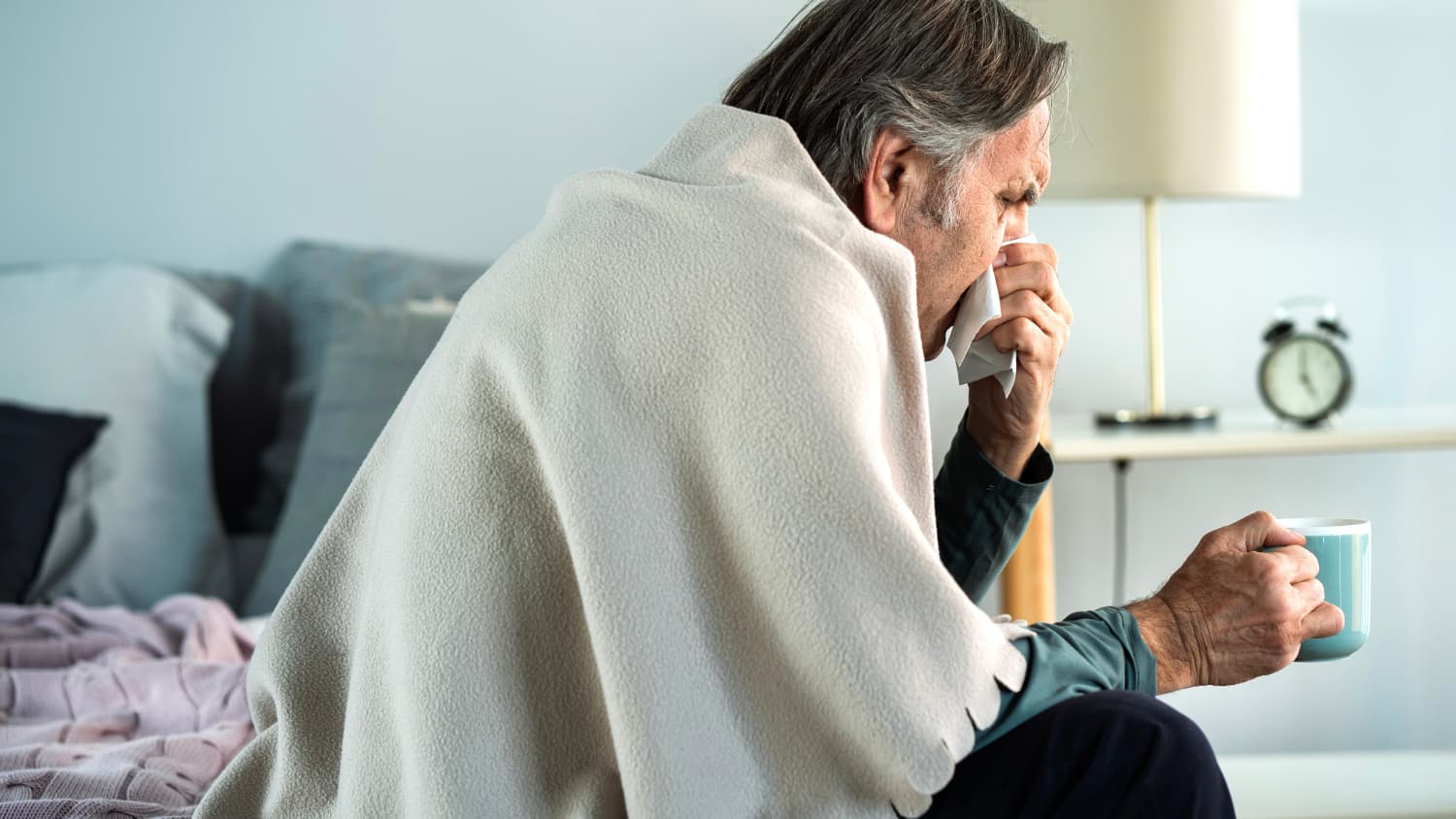 Ein erkälteter Mann mit Taschentuch und Teetasse sitzt auf seinem Bett