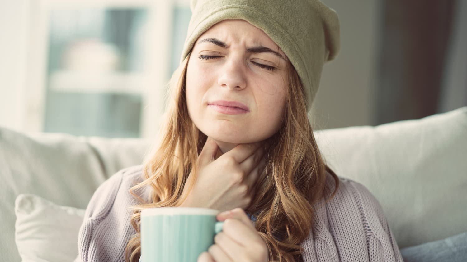 Eine Frau mit Mütze und Tasse langt sich an ihren schmerzenden Hals