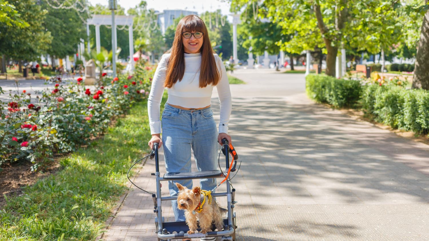 Eine junge Frau geht mit Hilfe einer Gehhilfe auf der ein Terrier sitzt in einem Park spazieren