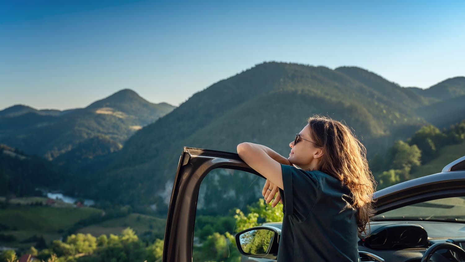 Eine Frau steht an ihrer Autotür gelehnt im Sommer mit Ausblick auf die Berge