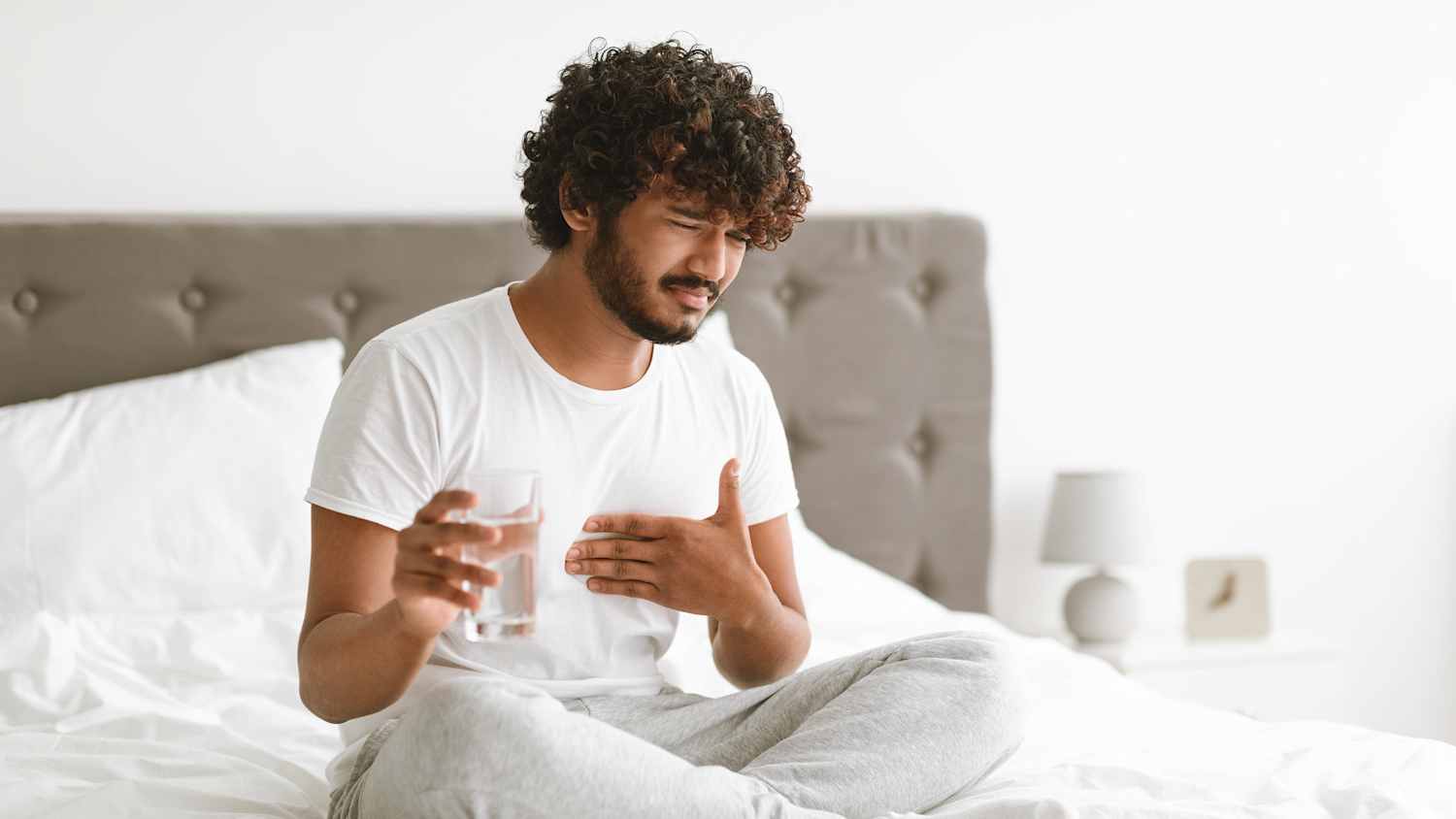 Ein Mann sitzt auf seinem Bett mit einem Glas Wasser in der Hand er hat offensichtlich Sodbrennen