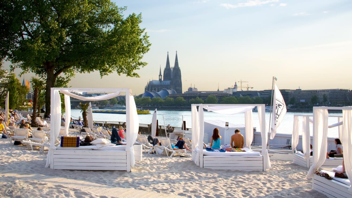 Der 689 Cologne Beach Club am Rheinufer in Köln mit weißen Beach Lounges und dem Kölner Dom im Hintergrund