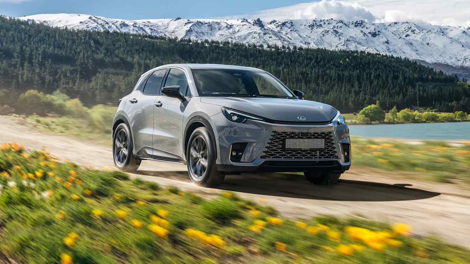 Front und Seitenansicht eines Lexus LBX der auf einer unasphaltierten Straße in Neuseeland fährt. Im Hintergrund ist ein See und Berge zu sehen