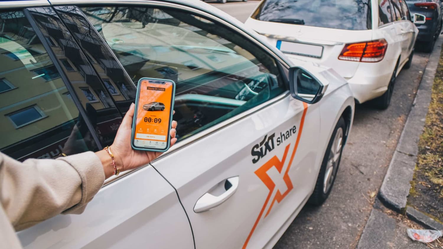Eine Frau hat die Sixt Share App auf ihrem Handy geöffnet und steht damit vor einem Carsharing Auto