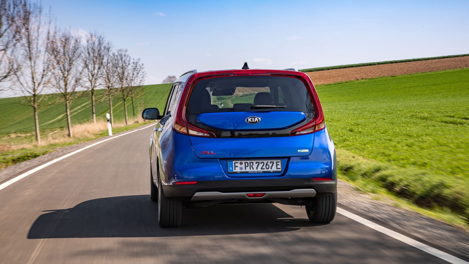 Blauer Kia E-Soul faehrt auf Strasse durch Landschaft