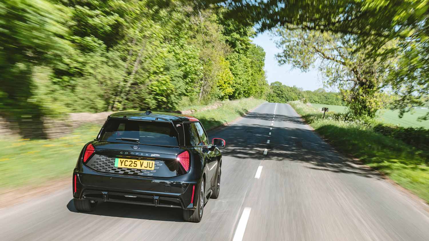 Der Mini John Cooper in  Electric Midnight Black von hinten fahrend auf einer Landstraße