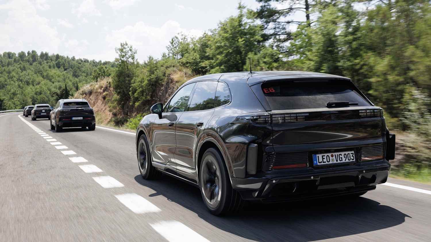 Heckansicht vier fahrender Porsche Cayenne E auf einer Straße mit Bäumen im Hintergrund