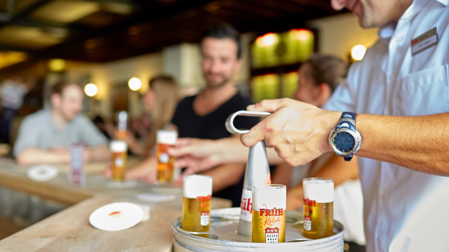 Ein Kellner bringt Gaesten ein frisch gezapttes Kölsch an den Tresen.