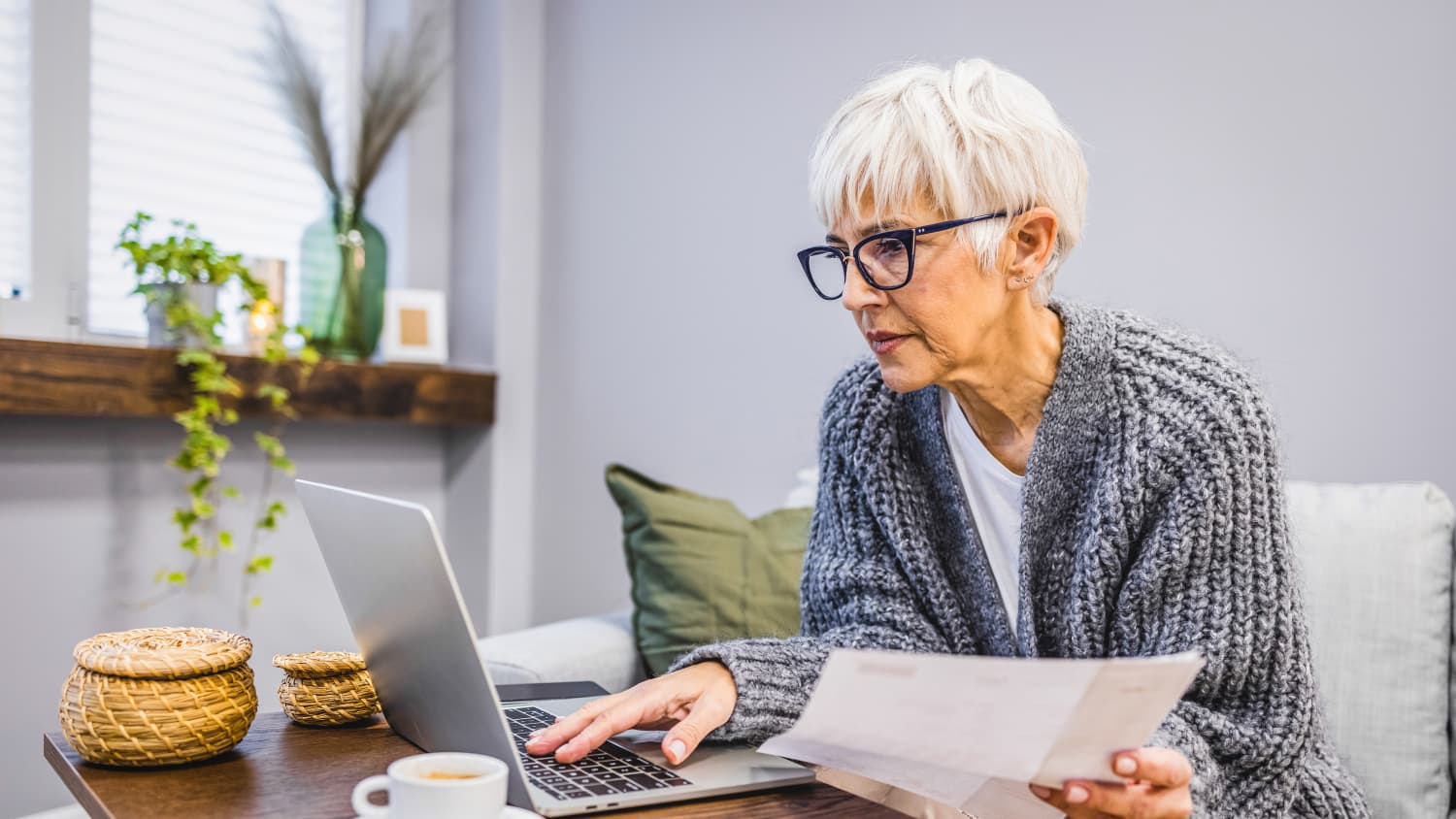 Eine ältere Dame sitzt mit einem Zettel in der Hand am Laptop