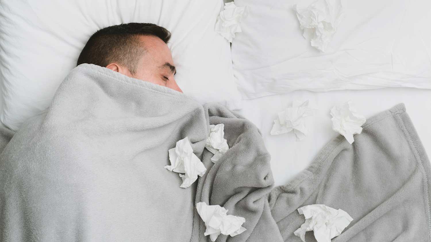 Ein Mann liegt mit vielen benutzen Taschentüchern krank im Bett