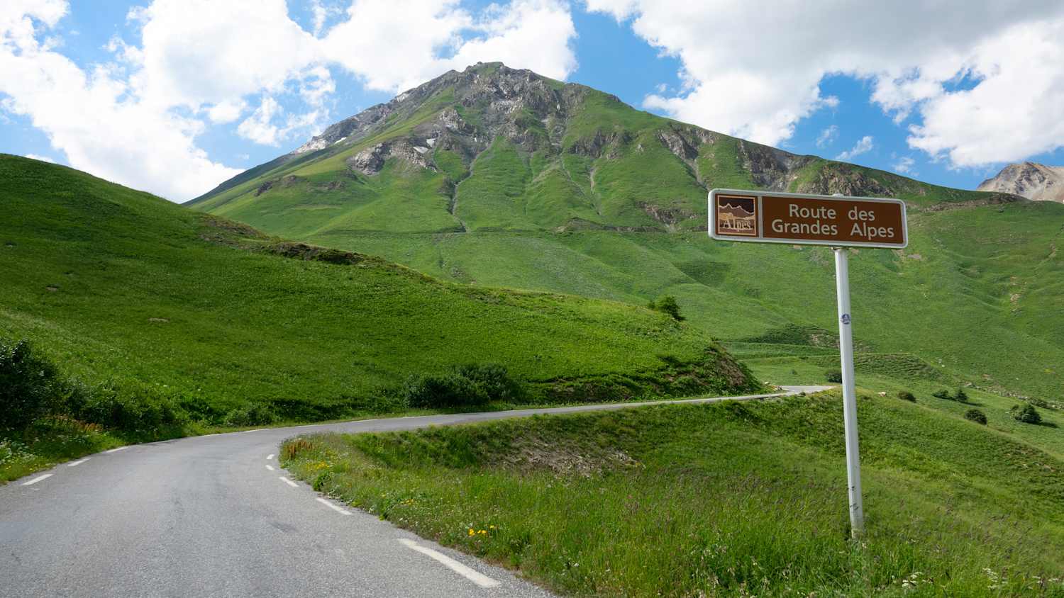 Eine leere Asphaltstraße windet sich um den grünen Hügel und vorbei an einem braunen Straßenschild, das Touristen darauf hinweist, dass sie auf der Route des Grandes Alpes unterwegs sind.