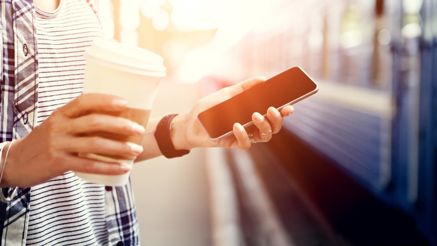 Eine Frau steht mit ihrem Smartphone und einen Coffee-to-go Becher vor einem Zug