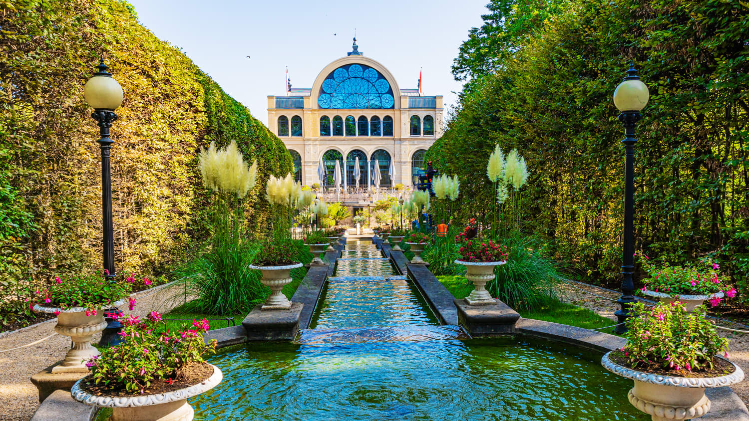 Botanischer Garten Köln mit seinem großen von Laternen und Pflanzen umsäumten Brunnen im Vordergrund im HIntergrund ist ein historisches Gebäude zu sehen