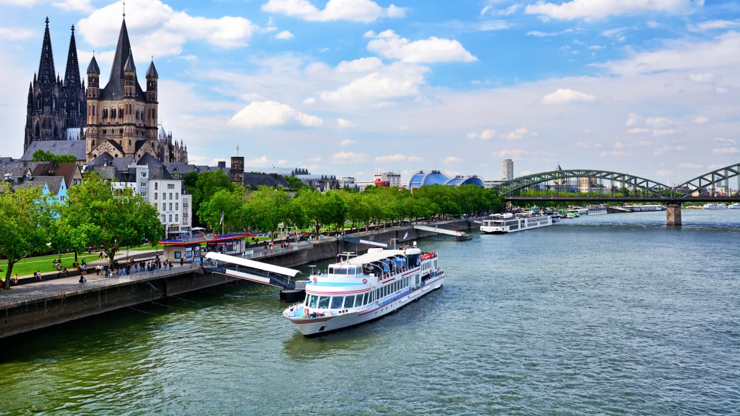 Ein Boot legt in Köln am Rhein an einem Bootssteg an, im Hintergrund sieht man die Hohenzollernbrücke und die Kirche St. Martin.