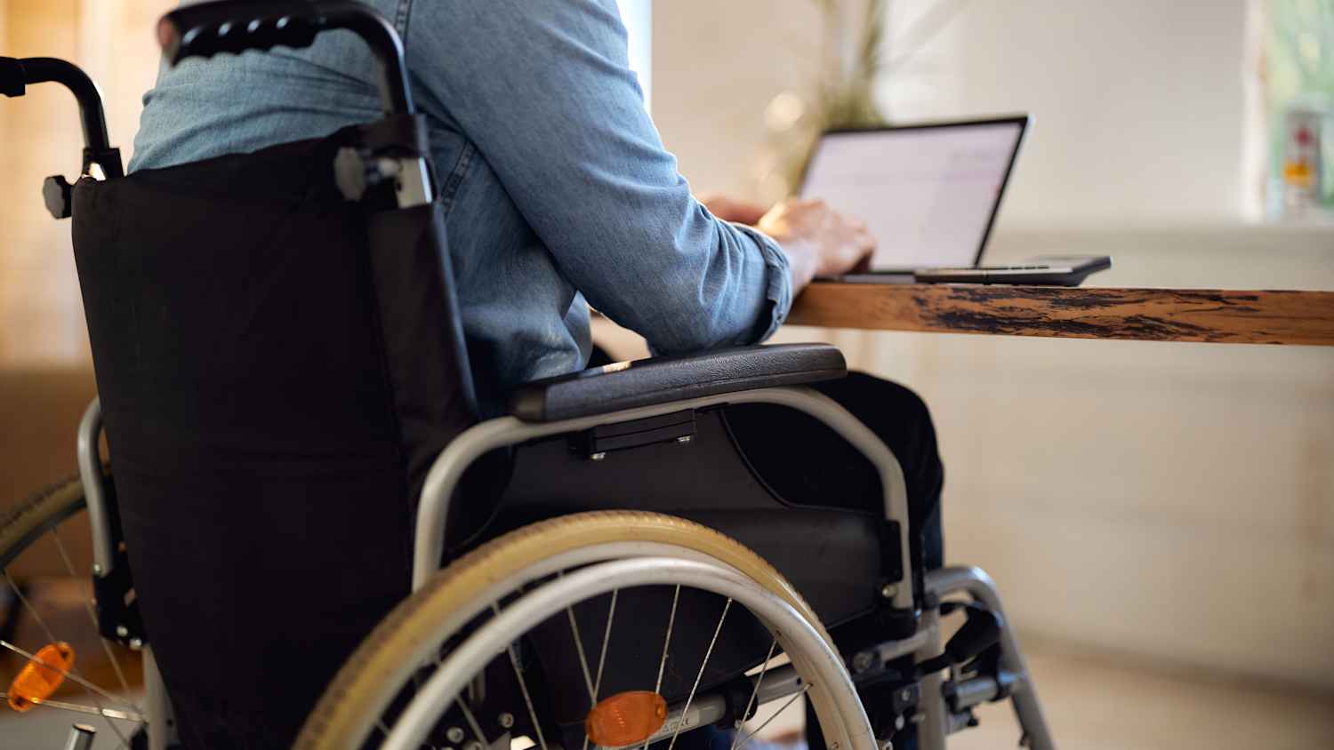 Ein Mann im Rollstuhl sitzt am Schreibtisch und schreibt am Laptop