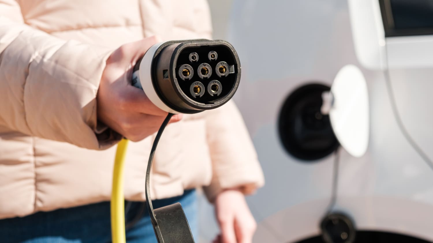 Eine Frau in heller Winterjacke hält den Ladestecker vom Auto in die Kamera