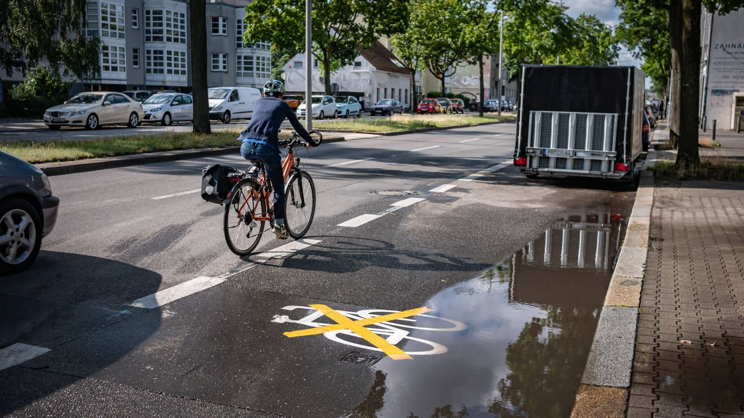 Ein Anhänger parkt auf einem durchgestrichenen Radweg während ein Auto und ein Fahrradfahrer auf der Straße fahren