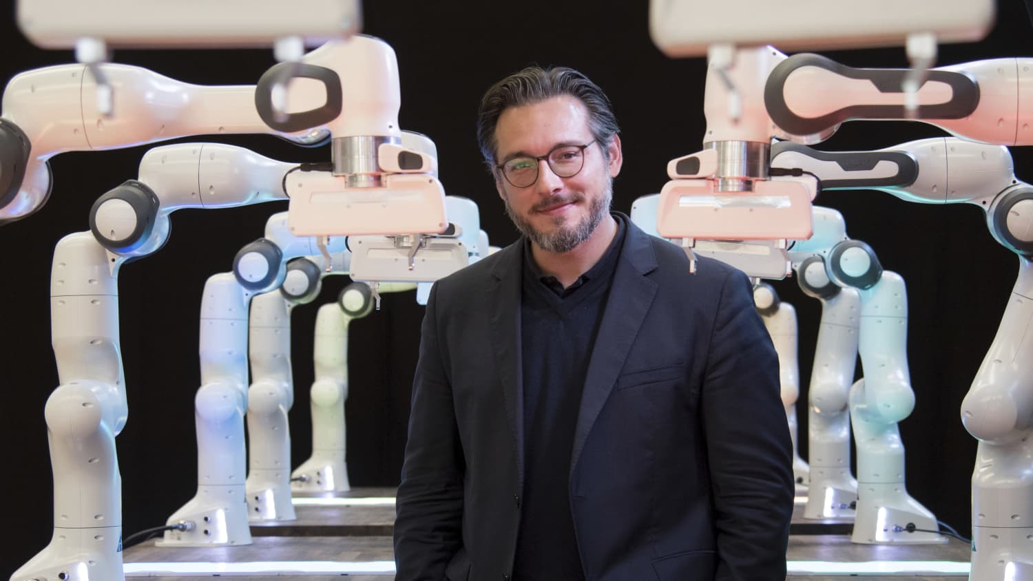 Professor Sami Haddadin im Porträt, viele Roboterarme sind im Hintergrund zu sehen